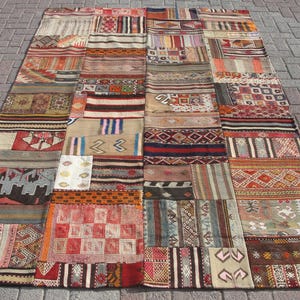 May include: A large, rectangular patchwork rug composed of various rectangular and square pieces. The rug features a mix of colors, including red, brown, beige, and blue, with geometric and striped patterns. The rug is laid out on a brick surface.