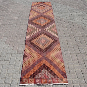 May include: A rectangular runner rug with a geometric diamond pattern in shades of orange, brown, red, and beige. The rug has a woven texture and a white fringe at the ends. The rug is placed on a brick surface.