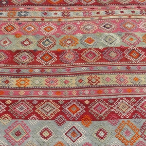 May include: A colorful, patterned rug with a geometric design. The rug features horizontal stripes in shades of red, pink, orange, and gray. The pattern includes diamond and square shapes. The rug is made of woven fabric.