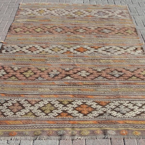 K&ouml;nnte beinhalten: Ein rechteckiger, handgewebter Teppich mit geometrischem Muster. Der Teppich weist ein sich wiederholendes Design aus Rauten und Streifen in Braun-, Orange-, Beige- und Gelbt&ouml;nen auf. Der Teppich hat einen Fransenrand und liegt auf einer Ziegeloberfl&auml;che.