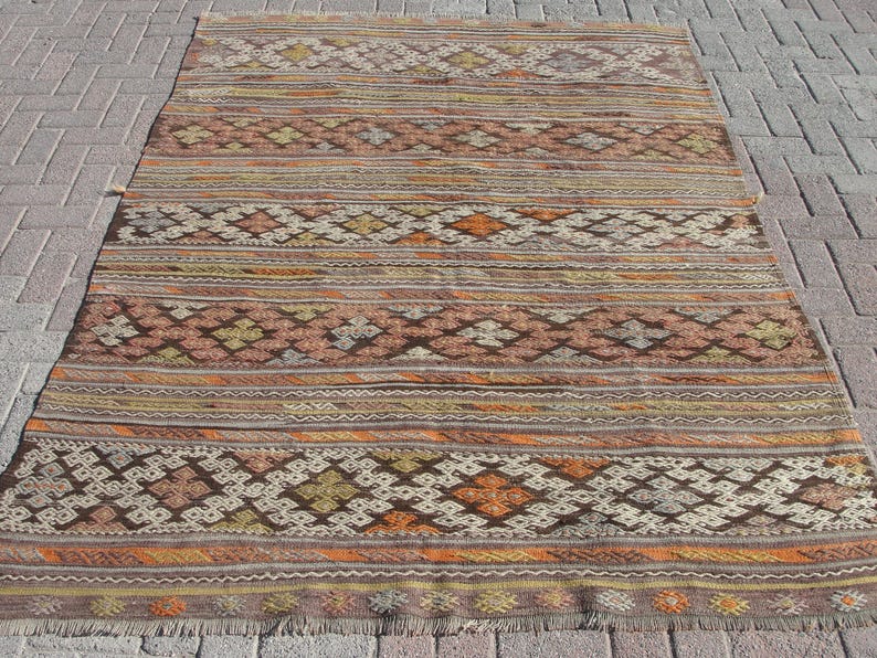 K&ouml;nnte beinhalten: Ein rechteckiger, handgewebter Teppich mit geometrischem Muster. Der Teppich weist ein sich wiederholendes Design aus Rauten und Streifen in Braun-, Orange-, Beige- und Gelbt&ouml;nen auf. Der Teppich hat einen Fransenrand und liegt auf einer Ziegeloberfl&auml;che.