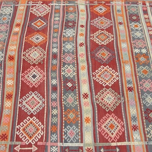 May include: A woven rug with a striped pattern in shades of red, gray, orange, and pink. The rug features geometric diamond and zig-zag designs. The rug's design is symmetrical, with vertical stripes of varying widths and colors.