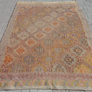 May include: A rectangular, vintage rug with a geometric pattern in shades of orange, yellow, and brown. The rug features diamond and star-like motifs, with a beige border and fringe. The rug is placed on a brick surface.