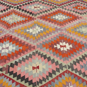 Op de afbeelding: Een kleurrijk tapijt met een geometrisch patroon in tinten rood, oranje, roze, grijs en wit.