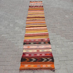 May include: A long, narrow, handwoven rug with a striped pattern in shades of orange, yellow, red, and brown. The rug features geometric designs at the ends and is likely made of natural fibers. This runner rug is suitable for a hallway or entryway.