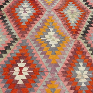 Op de afbeelding: Een kleurrijk tapijt met een geometrisch patroon in tinten roze, oranje, rood, grijs en wit. Het patroon bestaat uit herhalende driehoeken en ruiten.
