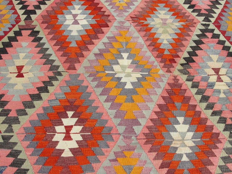 Op de afbeelding: Een kleurrijk tapijt met een geometrisch patroon in tinten roze, oranje, rood, grijs en wit. Het patroon bestaat uit herhalende driehoeken en ruiten.