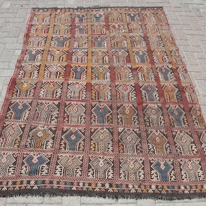 May include: A rectangular rug with a repeating geometric pattern in shades of brown, red, blue, and yellow. The rug has a dark brown fringe along the bottom edge. The pattern is composed of stylized tree-like shapes within a grid.
