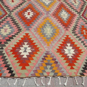 Op de afbeelding: Een traditioneel Turks kilim tapijt met een geometrisch patroon in tinten rood, oranje, geel, roze, grijs en bruin. Het tapijt heeft een franje rand.