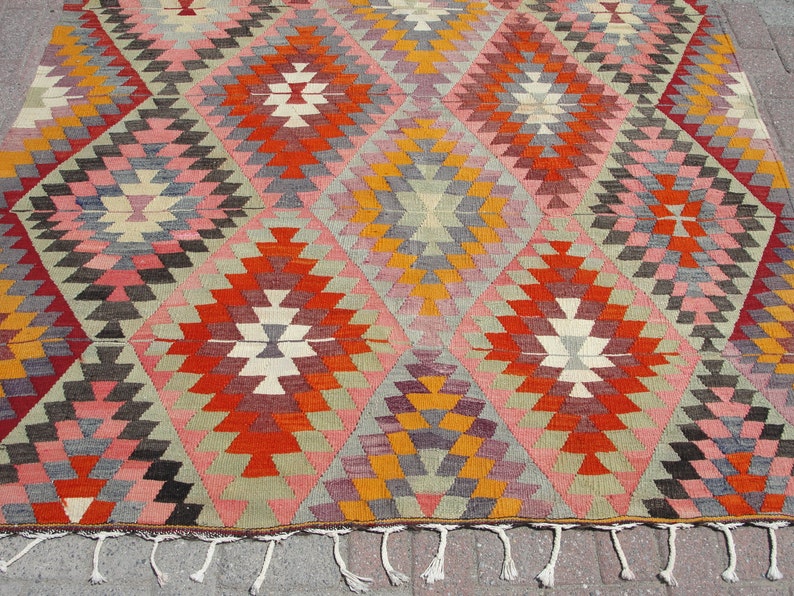 Op de afbeelding: Een traditioneel Turks kilim tapijt met een geometrisch patroon in tinten rood, oranje, geel, roze, grijs en bruin. Het tapijt heeft een franje rand.