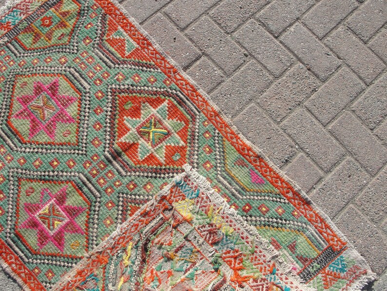 Puede incluir: Una alfombra colorida con patrones geom&eacute;tricos en tonos de verde, rojo, naranja y rosa. La alfombra tiene un borde con flecos y est&aacute; sobre una superficie de hormig&oacute;n gris.
