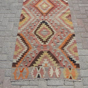 May include: A rectangular, patterned rug with a geometric design. The rug features a central diamond motif in shades of peach, brown, and orange, surrounded by zig-zag and triangular patterns. The rug is approximately 3 feet wide and 6 feet long.