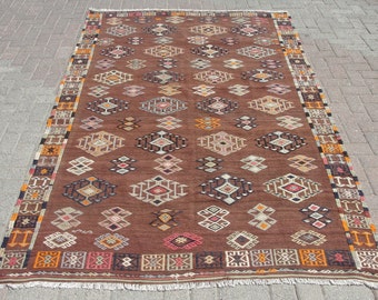 May include: A rectangular brown rug with an intricate geometric pattern. The rug features repeating diamond and geometric shapes in various colors, including white, red, and orange. The border has a detailed design.