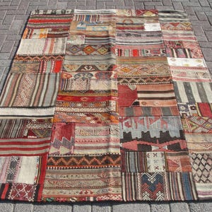 May include: A patchwork rug with a variety of patterns and colors, including red, brown, beige, and blue. The rug is rectangular and appears to be made of multiple pieces of fabric sewn together. The rug is placed on a gray brick surface.