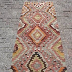 May include: A rectangular, patterned rug with a geometric design. The rug features a central diamond motif in shades of orange, red, and brown, surrounded by zig-zag patterns in beige, gray, and yellow. The rug is placed on a brick surface.