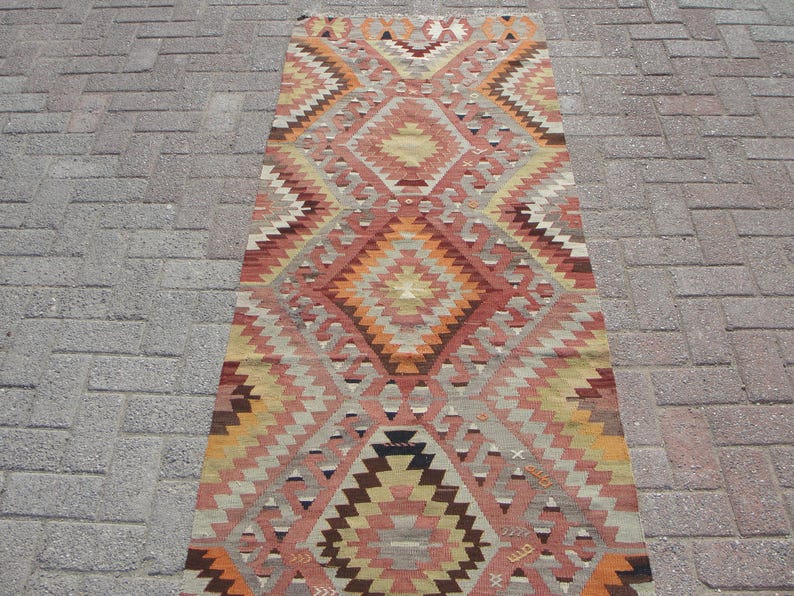 May include: A rectangular, patterned rug with a geometric design. The rug features a central diamond motif in shades of orange, red, and brown, surrounded by zig-zag patterns in beige, gray, and yellow. The rug is placed on a brick surface.
