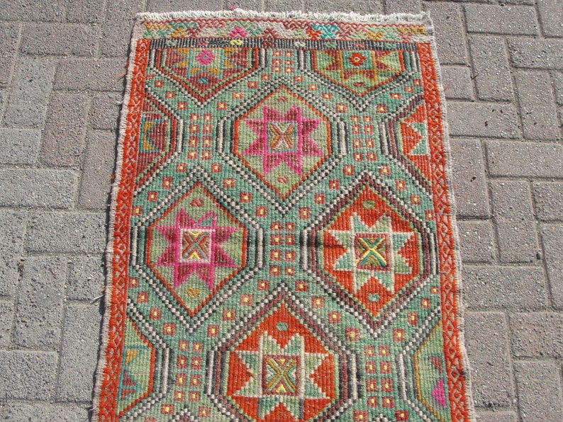 Puede incluir: Una alfombra turca vintage con un patr&oacute;n geom&eacute;trico en tonos de rojo, verde, naranja, rosa y amarillo. La alfombra tiene un borde con flecos y est&aacute; hecha de lana.