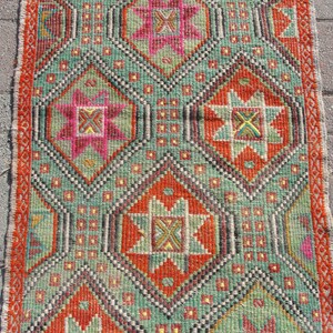 Puede incluir: Una alfombra de color verde, naranja, rosa y amarillo con un dise&ntilde;o geom&eacute;trico. La alfombra tiene un borde con flecos.