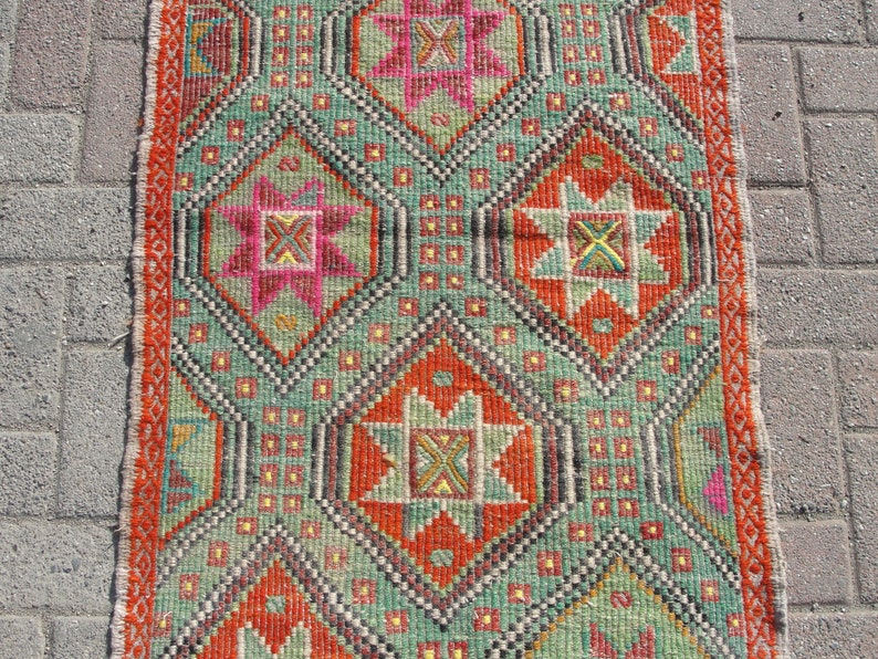 Puede incluir: Una alfombra de color verde, naranja, rosa y amarillo con un dise&ntilde;o geom&eacute;trico. La alfombra tiene un borde con flecos.