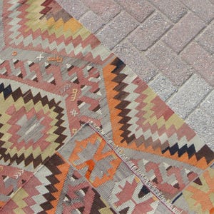 May include: A section of a colorful, patterned rug with geometric designs in shades of pink, orange, brown, and gray. The rug's edge is visible, showing the fringed border. The rug is placed on a brick surface.
