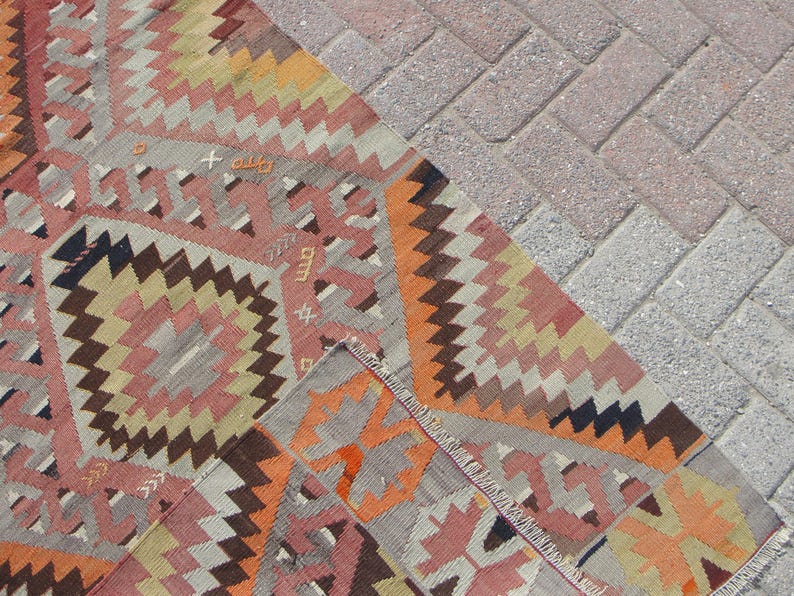 May include: A section of a colorful, patterned rug with geometric designs in shades of pink, orange, brown, and gray. The rug's edge is visible, showing the fringed border. The rug is placed on a brick surface.