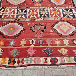 May include: A red, hand-woven rug with a geometric pattern in shades of blue, pink, orange, and white. The rug has a fringe border and is made of wool.