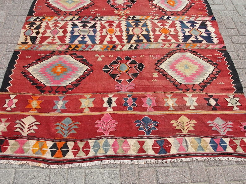 May include: A red, hand-woven rug with a geometric pattern in shades of blue, pink, orange, and white. The rug has a fringe border and is made of wool.