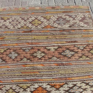 K&ouml;nnte beinhalten: Ein rechteckiger Teppich mit einem geometrischen Muster in Braun-, Orange- und Cremet&ouml;nen. Der Teppich weist horizontale Streifen und rautenf&ouml;rmige Motive auf. Der Teppich liegt auf einer Ziegeloberfl&auml;che.