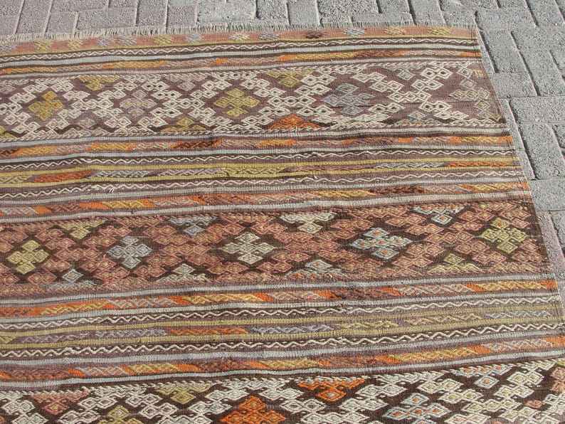 K&ouml;nnte beinhalten: Ein rechteckiger Teppich mit einem geometrischen Muster in Braun-, Orange- und Cremet&ouml;nen. Der Teppich weist horizontale Streifen und rautenf&ouml;rmige Motive auf. Der Teppich liegt auf einer Ziegeloberfl&auml;che.