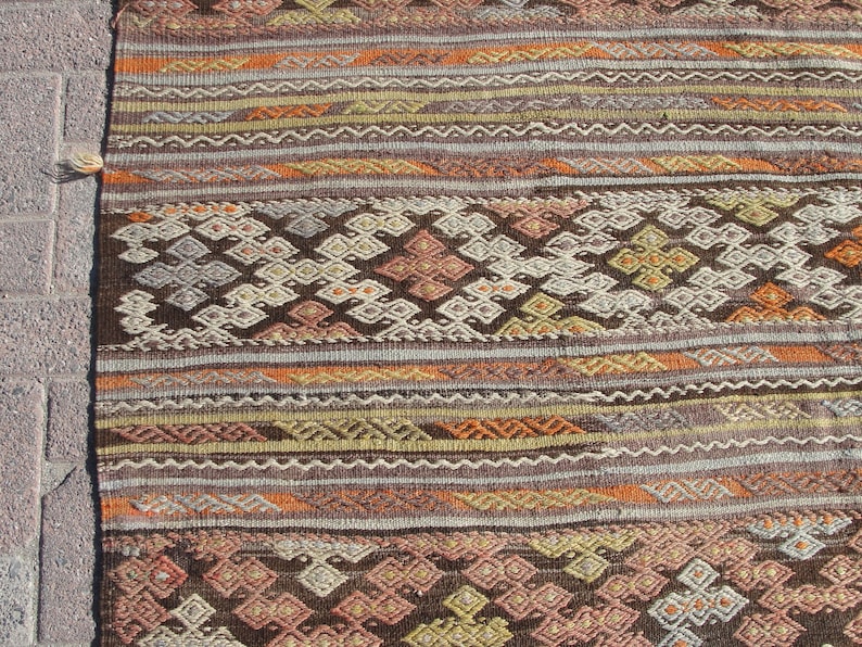K&ouml;nnte beinhalten: Nahaufnahme eines gewebten Teppichs mit geometrischem Muster. Der Teppich zeigt eine Mischung aus Farben, darunter Braun, Orange, Gelb und Grau. Das Design umfasst Diamanten und Streifen, die ein strukturiertes Aussehen erzeugen. Der Teppich ist wahrscheinlich aus Naturfasern gefertigt.