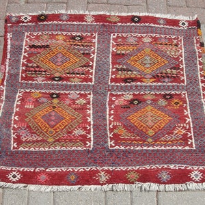 May include: A red, blue, and white patterned rug with a geometric design. The rug has a fringe border and is made of wool.