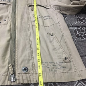 May include: Tan jacket with a hood, zipper closure, and multiple pockets. The jacket has text on the front and sleeve. The length of the jacket is approximately 27 inches.