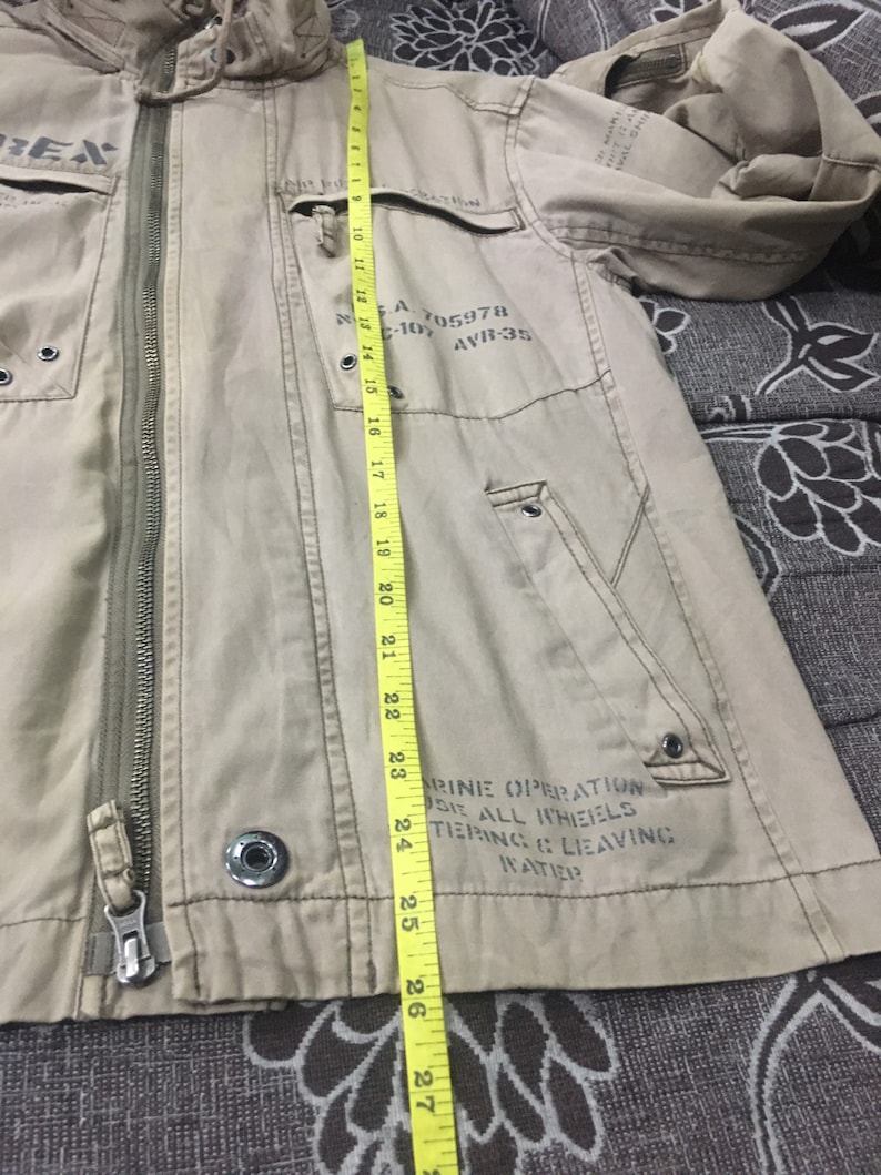 May include: Tan jacket with a hood, zipper closure, and multiple pockets. The jacket has text on the front and sleeve. The length of the jacket is approximately 27 inches.