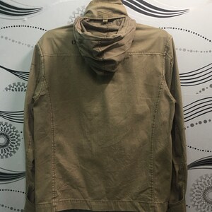 May include: A khaki-colored jacket with a hood, hanging against a patterned background. The jacket has a full zipper, pockets, and a buttoned cuff. The hood is folded down. The jacket appears to be made of a durable fabric.