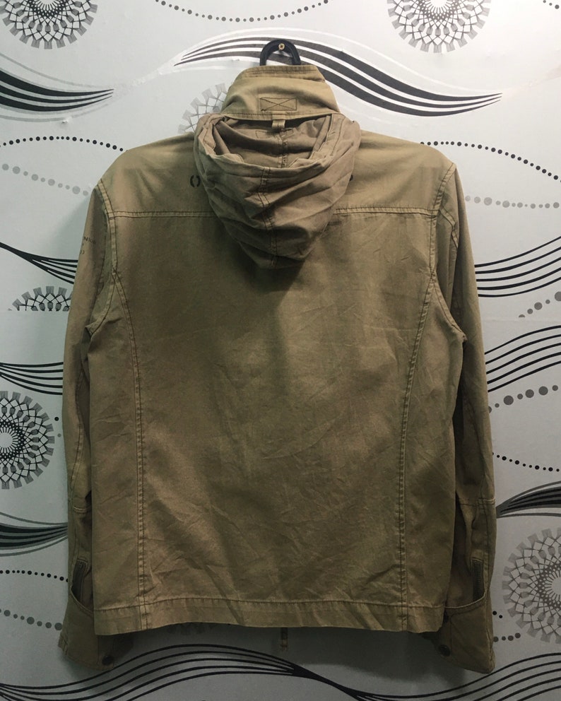 May include: A khaki-colored jacket with a hood, hanging against a patterned background. The jacket has a full zipper, pockets, and a buttoned cuff. The hood is folded down. The jacket appears to be made of a durable fabric.