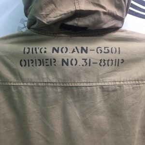 May include: Close-up of a khaki-colored jacket with a hood. The back of the jacket features stenciled black text: "DIVG NO.AN-6501" and "ORDER NO.31-8011P". The jacket has a simple design with a visible seam across the upper back.