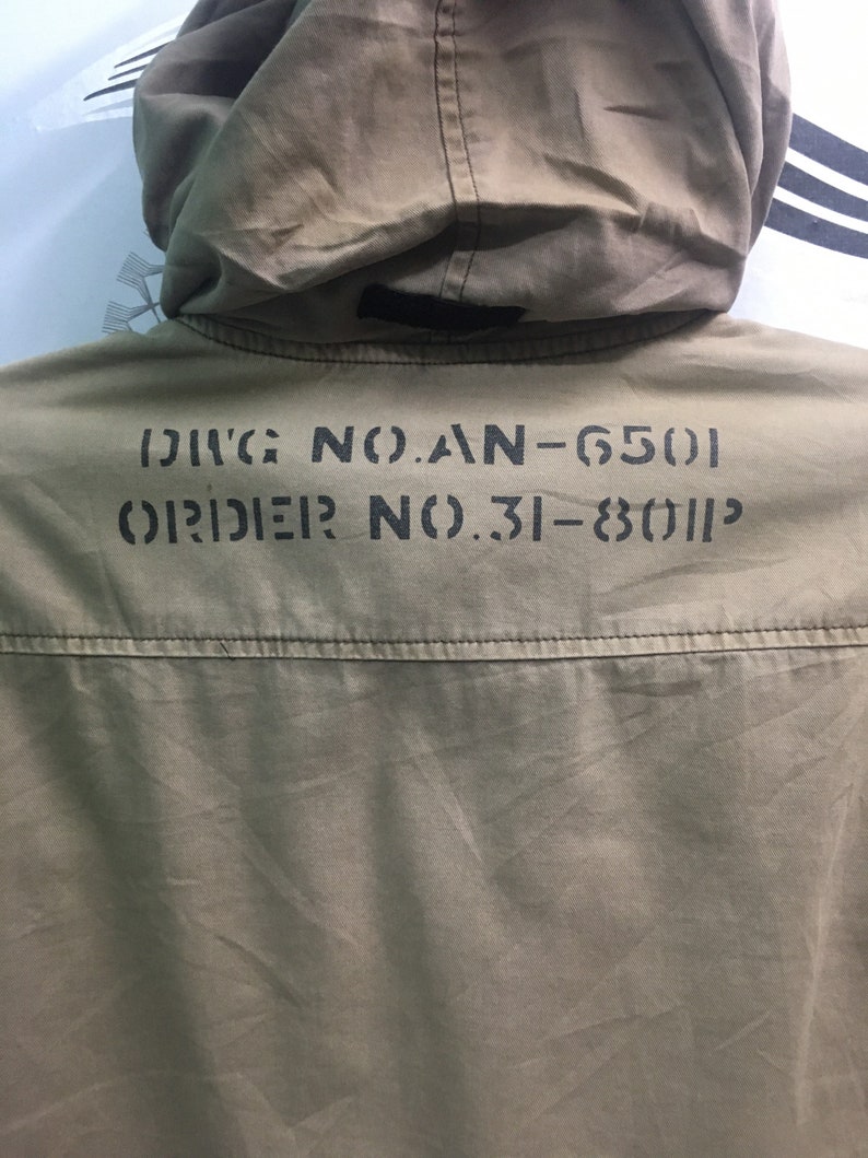 May include: Close-up of a khaki-colored jacket with a hood. The back of the jacket features stenciled black text: "DIVG NO.AN-6501" and "ORDER NO.31-8011P". The jacket has a simple design with a visible seam across the upper back.