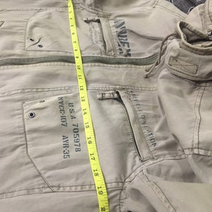 May include: Tan jacket with multiple pockets and a zipper closure. The jacket features printed text and a hood. A yellow measuring tape shows the jacket's length is approximately 21 inches.