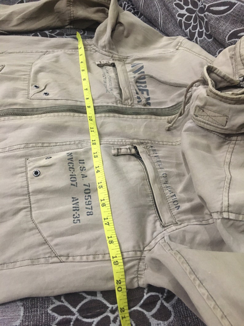 May include: Tan jacket with multiple pockets and a zipper closure. The jacket features printed text and a hood. A yellow measuring tape shows the jacket's length is approximately 21 inches.