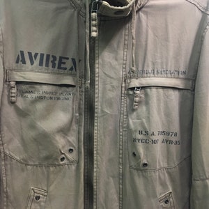 May include: Khaki-colored jacket with the word "AVIREX" printed in black on the front. The jacket has multiple pockets, a zipper closure, and text that reads "FLAME & POWER PLANT RADIAL & PISTON ENGINE" and "U.S.A. 705978".