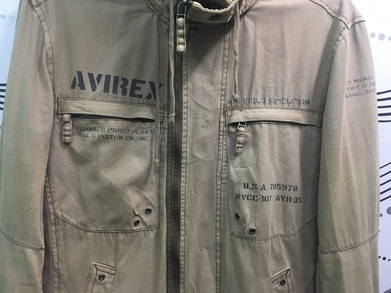 May include: Khaki-colored jacket with the word "AVIREX" printed in black on the front. The jacket has multiple pockets, a zipper closure, and text that reads "FLAME & POWER PLANT RADIAL & PISTON ENGINE" and "U.S.A. 705978".