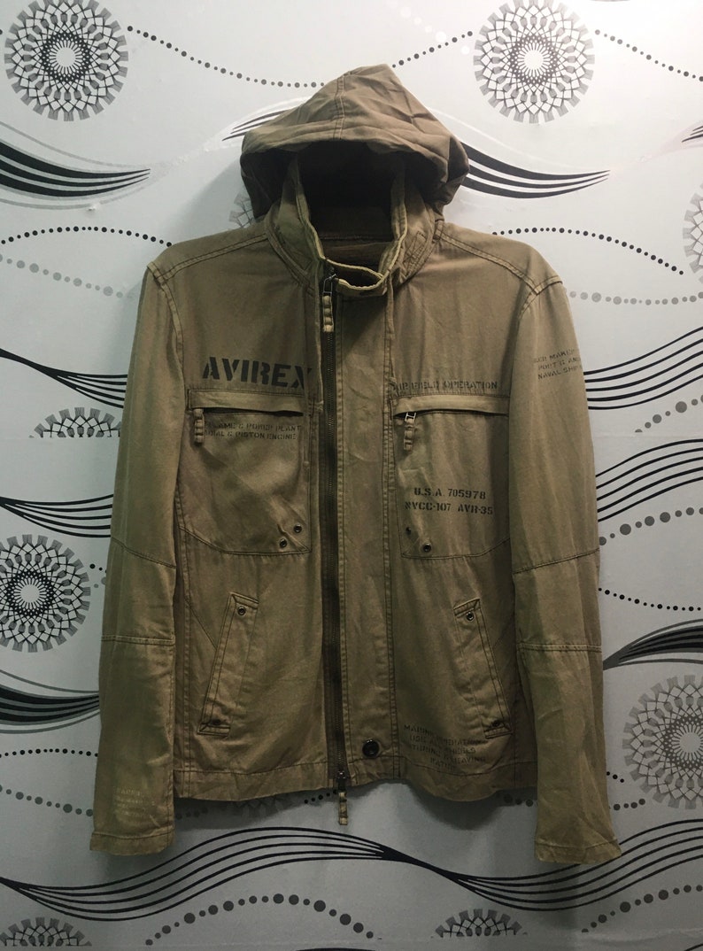 May include: A tan jacket with a hood and a full-length zipper. The jacket has multiple pockets and the word "AVIREX" printed on the chest. Additional text and graphics are printed on the jacket, including "U.S.A. 705978".