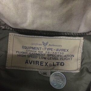 May include: Close-up of an olive green jacket label with the text "EQUIPMENT-TYPE-AVIREX" and "AVIREX LTD". The label also includes the stock number 28-A6755 and the size "M". A coin is visible.