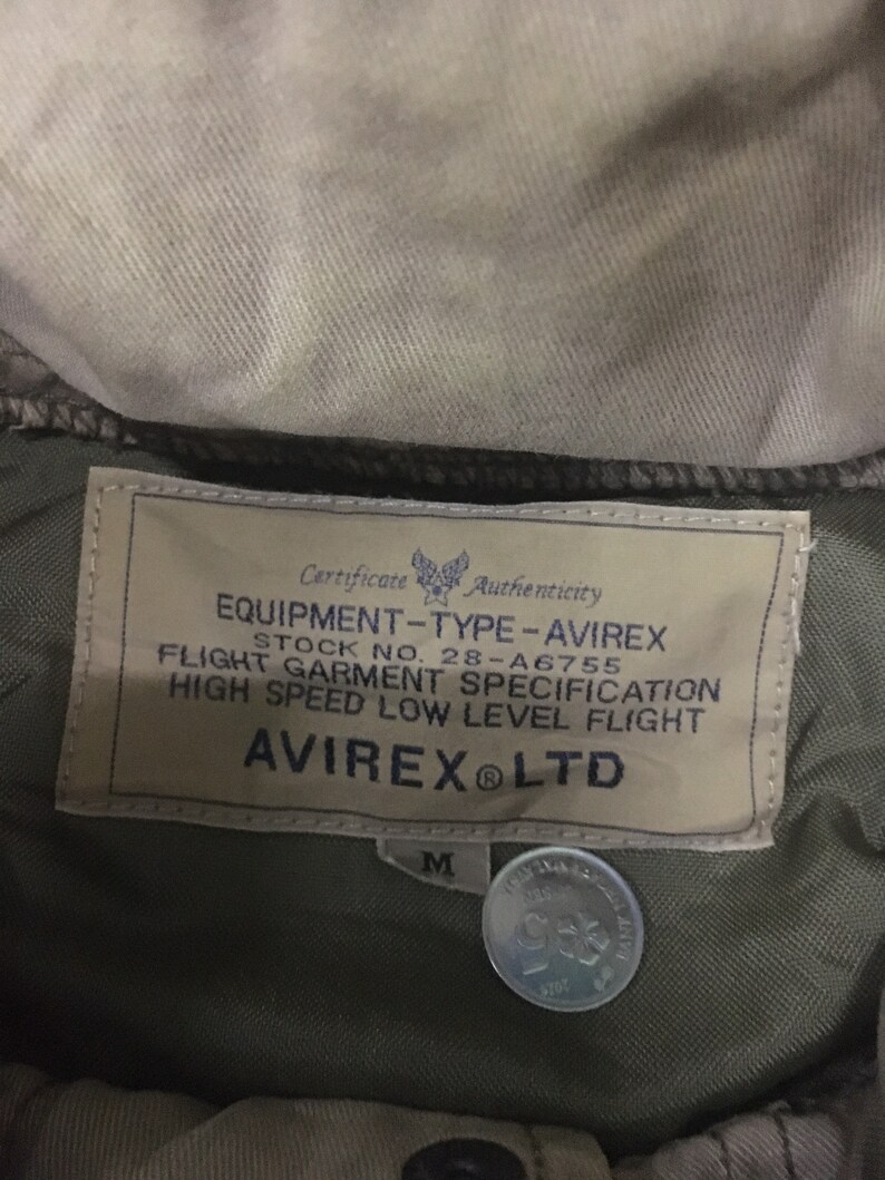 May include: Close-up of an olive green jacket label with the text "EQUIPMENT-TYPE-AVIREX" and "AVIREX LTD". The label also includes the stock number 28-A6755 and the size "M". A coin is visible.