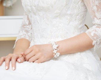 May include: A white bridal bracelet with delicate white flowers and silver accents. The bracelet is worn on the wrist of a person wearing a white lace dress.