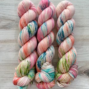 May include: Three skeins of hand-dyed yarn in a soft, pastel colour palette. The yarn features a blend of pink, peach, teal, and green hues, creating a variegated effect. The yarn is arranged on a light wood surface.