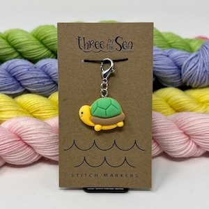 May include: A turtle-shaped stitch marker with a green shell, yellow body, and brown trim hangs from a silver clasp. The marker is attached to a brown card with the text "Three by the Sea designs" and "STITCH MARKERS". Colorful yarn skeins are in the background.