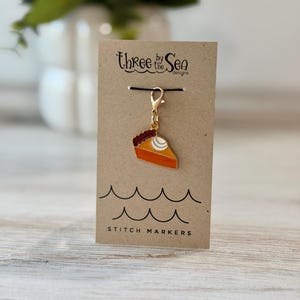 May include: A gold-toned metal stitch marker charm shaped like a slice of pumpkin pie with orange filling, a dark brown crust, and a white dollop of whipped cream, attached to a tan card labeled Three by the Sea Designs Stitch Markers.