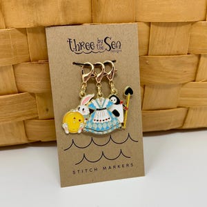 Op de afbeelding: Drie steekmarkeerders met Alice in Wonderland-figuren. De markeerders zijn goudkleurig en tonen een wit konijn met een zakhorloge, Alice in haar blauw-witte jurk en een pinguïn die een speelkaart vasthoudt. De kaart heeft de tekst "Three by the Sea Designs" en "Stitch Markers".