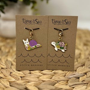 May include: Two stitch markers on brown cards. One is a white snail with a purple shell and floral accents. The other is a white turtle with floral accents. Each card has the text "Three by the Sea designs" and "Stitch Markers".